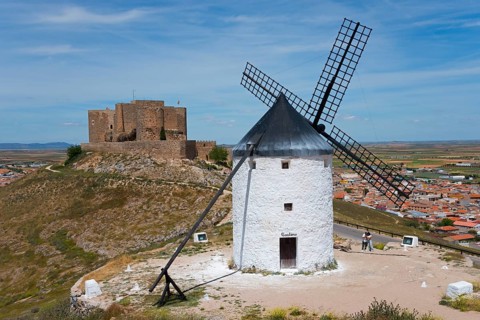 An old windmill in front of a medieval castle on a hill with a village in the background under a
