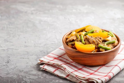 Fried pork with peaches, cashew and green beans in a wooden bowl on a gray concrete background.