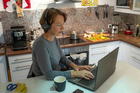 Woman, mid-50s, works from home, with laptop and communicates with colleagues via headset, home