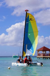 People on a colourful sailing boat in the clear sea near a jetty with a pavilion under a blue sky,