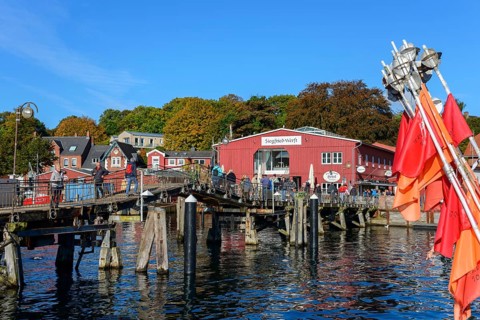 Sunlit harbour with boats and moorings in front of small colourful buildings, wooden bridge and