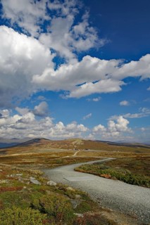 A hiking trail leads through a wide, hilly landscape towards the mountains under a blue sky,