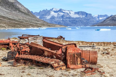 Rusty cars and barrels on a fjord in front of steep mountains, remains of a US airbase from the