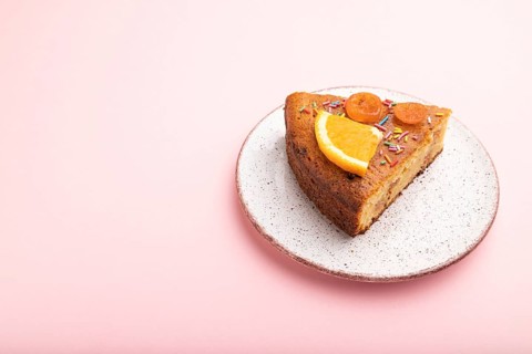 A piece of orange cake on a pastel pink background. Side view, copy space, close up