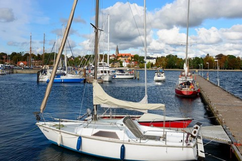 Sailboats lying in the calm waters of a marina, with a city view and cloudy sky in the background,