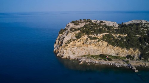 A drone shot shows a remote coastal strip with steep cliffs at the edge of the clear blue sea,