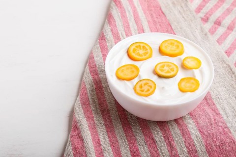 Greek yogurt with kumquat pieces in a white plate on a white wooden background, copy space