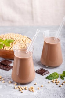 Organic non dairy soy chocolate milk in glass and wooden plate with soybeans on a gray concrete