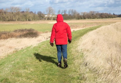 Woman wearing red winter jacket walking on raised coastal flood defence dyke embankment, Hollesley
