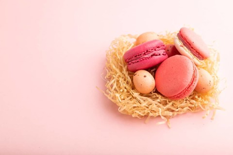 Purple and pink macaroons and chocolate eggs in decorative paper nest on pink pastel background.