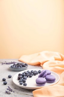 Purple macarons or macaroons cakes with blueberries on white ceramic plate on a gray and orange