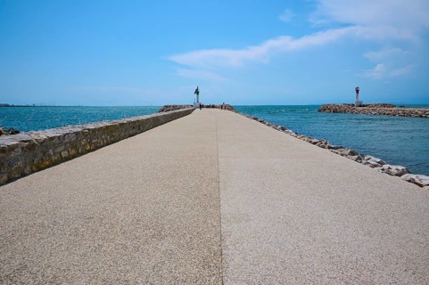 A long concrete path leads to the sea, flanked by breakwaters under a sunny, cloudy sky, summer, Le