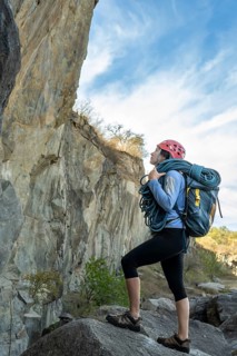 Female climber standing in front of a rock, carrying climbing rope, looking up, observing rocky