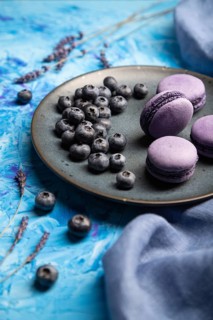 Purple macarons or macaroons cakes with blueberries on ceramic plate on a blue concrete background
