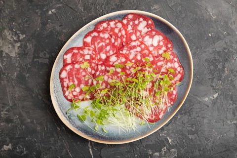 Slices of smoked cervelat salami sausage with spinach microgreen on black concrete background. Top