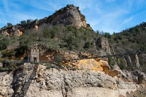 A rocky mountain landscape with dense vegetation under a bright blue sky, suspension bridge,