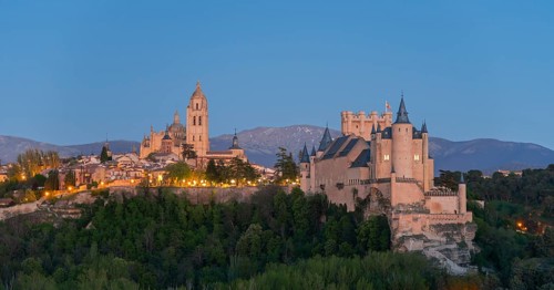 A majestic castle on a hill at dusk with illuminated buildings and a mountain backdrop, Cathedral,