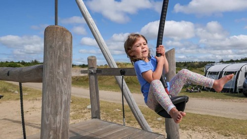 A child happily jumps on a zip wire in a wide, sunny playground setting with a cloudy, blue sky