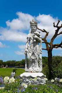 A stone Buddha statue surrounded by trees and flowers under a blue sky with clouds, Bacalhôa,