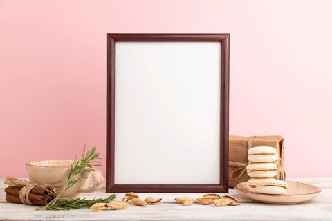Brown wooden frame mockup with cup of coffee, almonds and macaroons on pink pastel background.