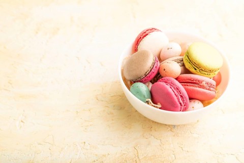 Multicolored macaroons and chocolate eggs in ceramic bowl on beige concrete background. side view,