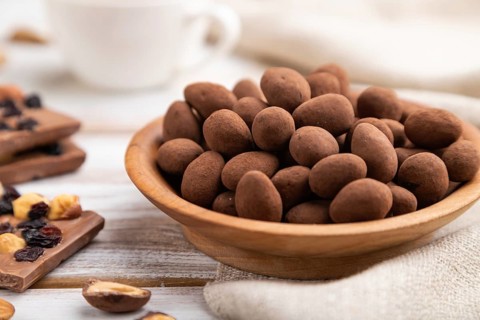 Almond in chocolate dragees in wooden plate and a cup of coffee on white wooden background and