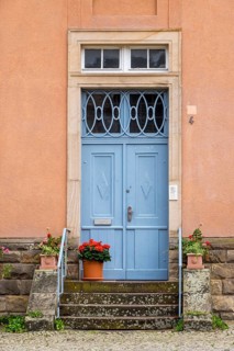 A blue front door with iron decorations on a pink façade, surrounded by potted plants,