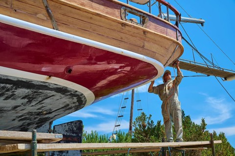A worker works on the red hull of a wooden sailing ship in the dry dock, Patmos Marine, boatyard,