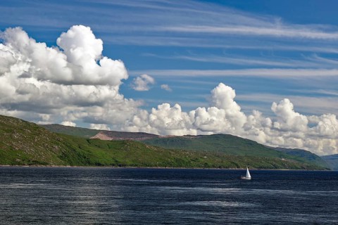 Single sailing boat on the sea, surrounded by a mountain landscape, under a sky with many clouds,