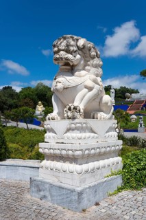 A stone lion statue on a pedestal, surrounded by plants under a blue sky with clouds, Bacalhôa,