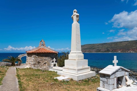 A tall monument next to a chapel in a cemetery with a view of the sea and surrounding mountains,