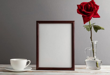 Brown wooden frame mockup with red rose in glass and white coffee cup on gray paper background.