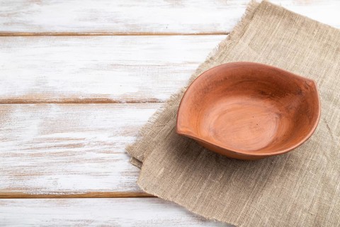 Empty clay brown bowl on white wooden background and linen textile. Side view, close up, copy space