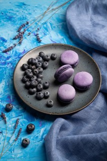 Purple macarons or macaroons cakes with blueberries on ceramic plate on a blue concrete background
