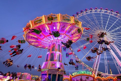 A lively funfair at dusk, illuminated by a large Ferris wheel and a colourful chain carousel,