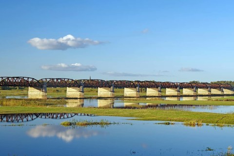 Long bridge over a calm river with reflections and green banks under a clear blue sky, autumn,
