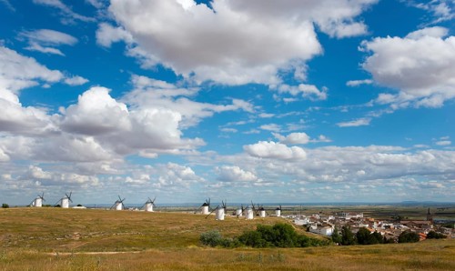 Wide landscape with windmills and a village in the background. The sky is blue with many white
