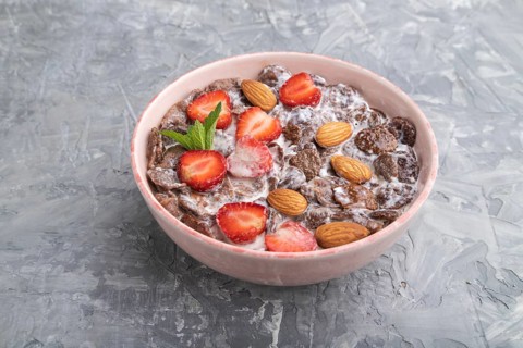Chocolate cornflakes with milk, strawberry and almonds in ceramic bowl on gray concrete background.