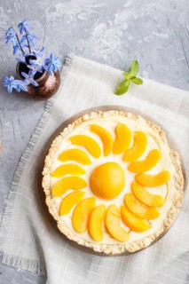 Round peach cheesecake and ceramic vase with blue flowers on a linen napkin on a gray concrete