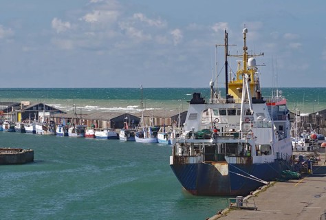 A large fishing boat moored at the harbour, with small fishing boats in the distance and the sea in