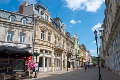 A lively city street with historic buildings under a blue sky on a sunny day, pedestrianised