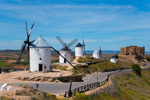 Several white windmills on a hill, with a castle in the background under a blue sky, Consuegra,