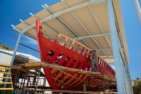 A partially built red wooden ship in a covered shipyard with a worker and blue sky, Patmos Marine,