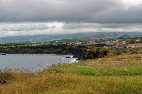 A village on the coast with green cliffs and a cloudy sky, Fenais da Luz, Sao Miguel Island,