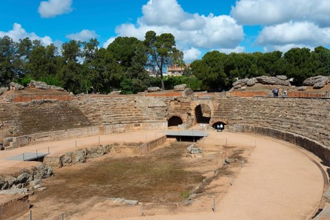 Partial view of a Roman amphitheatre with sandy floor and surrounding trees under a blue sky,