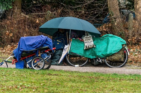 Homeless man in camp on park bench under umbrella with blanket and sleeping bag, bicycle covered