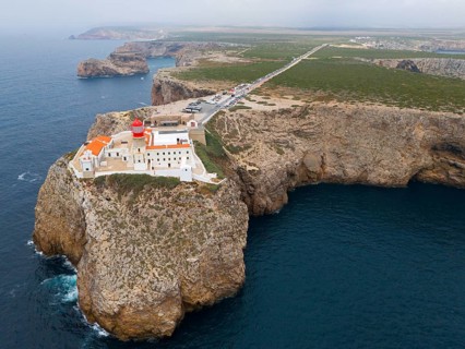 A remote lighthouse and several buildings stand on a cliff, with a wide view of the ocean and the