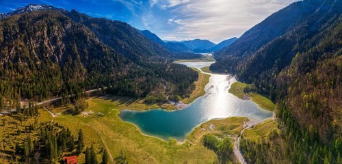 Mountain landscape with lake and forest, reflection of the sun in the water under a blue sky,