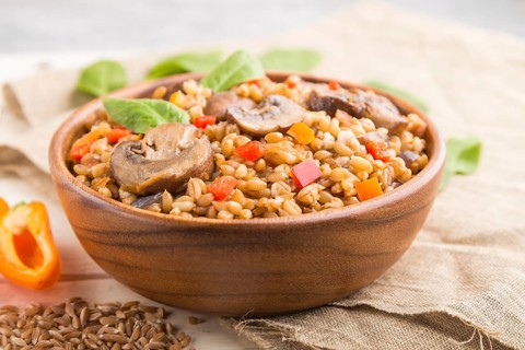 Spelt (dinkel wheat) porridge with vegetables and mushrooms in wooden bowl on a white wooden