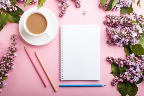 Purple lilac flowers and a cup of coffee with notebook and colored pencils on pastel pink